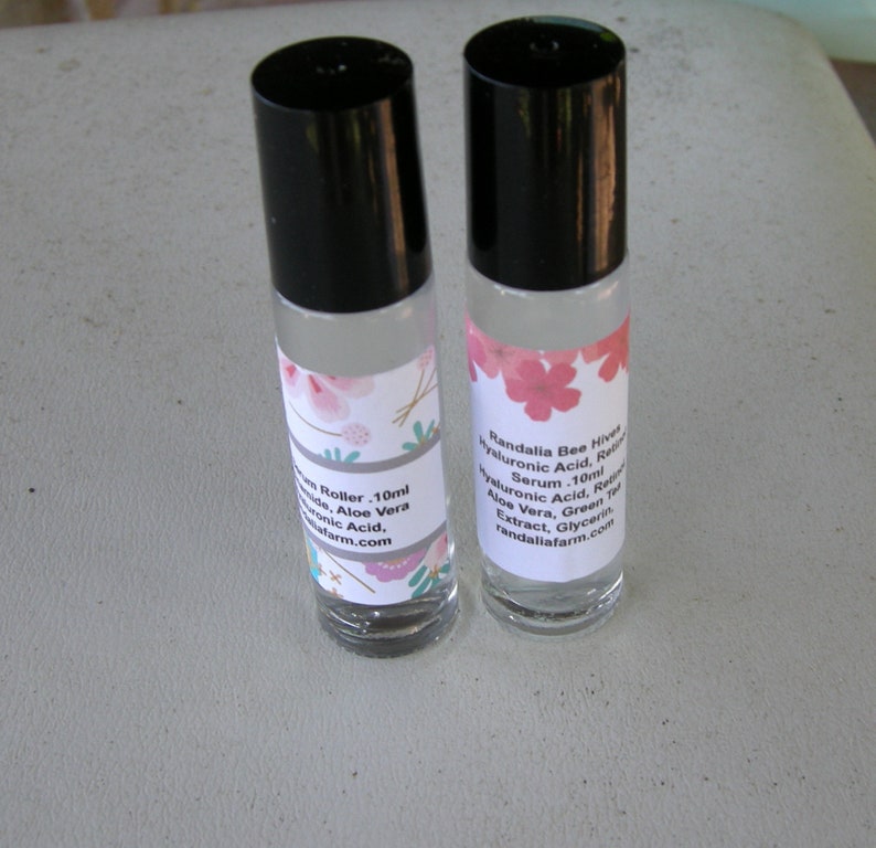 May include: Two clear glass roller bottles with black caps and labels. The labels are white with pink flowers and text that reads "Randalia Bee Hives Hyaluronic Acid, Retinol Serum - 10ml Hyaluronic Acid, Retinol, Aloe Vera, Green Tea Extract, Glycerin, randaliafarm.com".