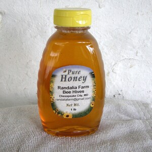 May include: A clear glass bottle of honey with a yellow lid. The label reads "Pure Honey", "Randalia Farm Bee Hives", "Chesapeake City, MD", "randaliafarm@gmail.com", and "Net Wt. 1 lb".
