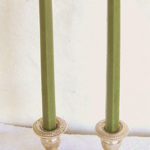 Puede incluir: Dos candelabros plateados con velas verdes. Los candelabros tienen una base cuadrada y un diseño detallado. Las velas son altas y rectangulares. El fondo es una pared blanca.