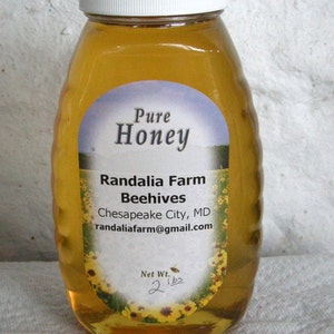 May include: A clear glass bottle of honey with a white lid and a label that reads "Pure Honey, Randalia Farm Beehives, Chesapeake City, MD, randaliafarm@gmail.com, Net Wt. 2 lbs".
