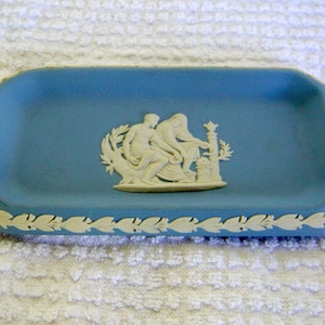 May include: A light blue ceramic trinket tray with a white raised design of two figures and a column. The tray has a white leaf border.