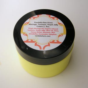 May include: A yellow jar with a black lid and a white label that reads "Randalia Bee Hives Moringa, Turmeric, Royal Jelly Cream 2, 4oz. Virgin Moringa Oil, Organic Infused Turmeric, Royal Jelly, Olive Oil, Shea, Cocoa Butter, Beeswax, Wax Emulsifier, Rosewater, Optiphen randaliafarm.com"
