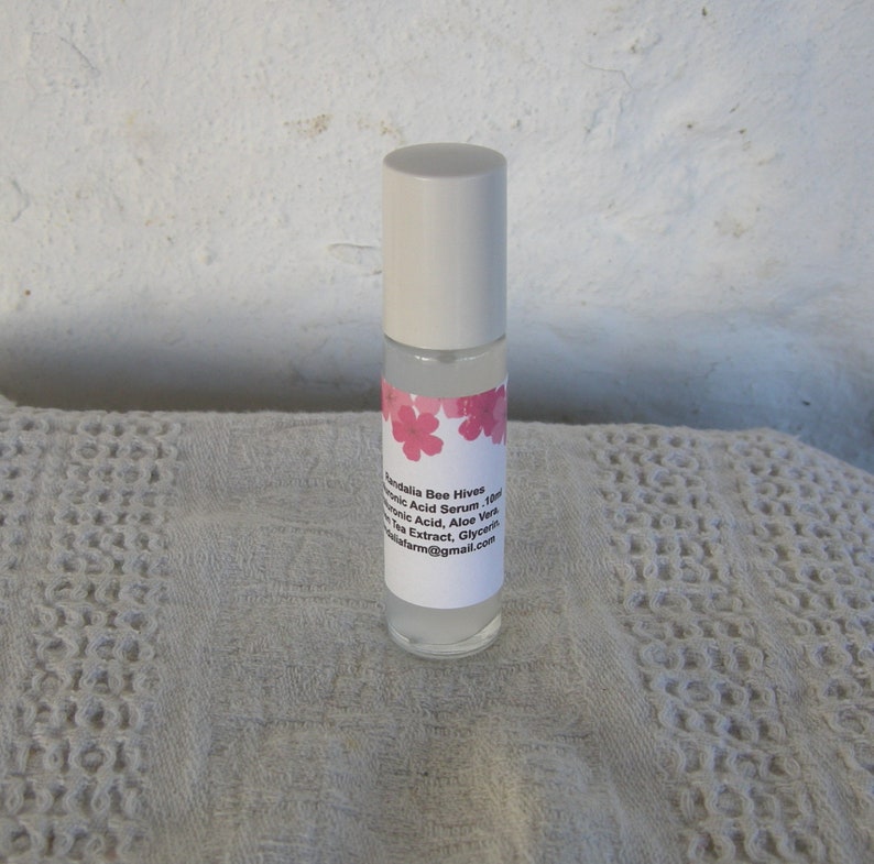 May include: A white roll-on bottle with a pink floral label that reads "Sandalia Bee Hives Hyaluronic Acid Serum 10ml", "Hyaluronic Acid, Aloe Vera, Green Tea Extract, Glycerin", and "sandaliafarm@gmail.com".