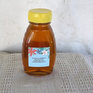 Puede incluir: Una botella de vidrio de miel con una tapa amarilla y una etiqueta que dice "Pine Barrens Wildflower Raw Honey 8oz. Randalia Bee Hives randaliafarm@gmail.com".