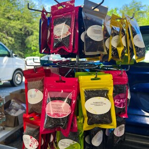 May include: A rotating metal display rack holds multiple small, resealable plastic pouches filled with various loose-leaf teas. The pouches come in vibrant colors including red, pink, yellow, and black, each featuring a white oval label with text.