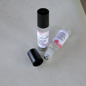 May include: Two clear glass roller bottles with black caps and metal roller balls. The bottles are filled with a clear liquid and have labels with floral designs and text that reads "Natural Essential Oil Blend, Aloe Vera, Hyaluronic Acid, Vitamin E Oil,  Nourishing Your Skin,  [website address]".