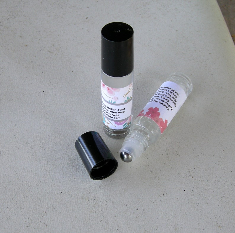May include: Two clear glass roller bottles with black caps and metal roller balls. The bottles are filled with a clear liquid and have labels with floral designs and text that reads "Natural Essential Oil Blend, Aloe Vera, Hyaluronic Acid, Vitamin E Oil,  Nourishing Your Skin,  [website address]".