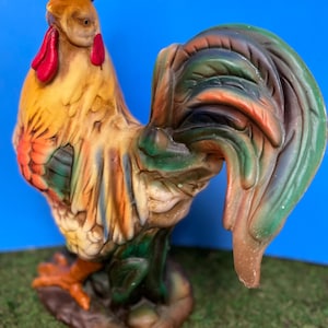 May include: A colorful ceramic rooster figurine with a red comb and wattle. The rooster has a green, brown, and orange tail.