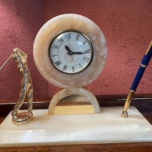 May include: A decorative desk clock with a round face and Roman numerals, set on a rectangular base. A gold-colored golfer figurine and a blue and gold pen are on either side of the clock.