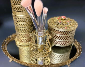 May include: A gold-toned vanity set with a round mirrored tray, two ornate containers with lids, and a decorative stand holding three makeup brushes.