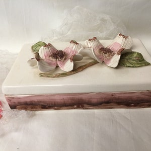 May include: A white ceramic trinket box with a pink and brown floral design. The box has a hinged lid and is decorated with three pink flowers with green leaves.