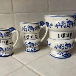 May include: Three white ceramic measuring pitchers with blue floral patterns. The pitchers are labeled 1/3 Cup, 1/2 Cup, and 1 Cup in black gothic-style lettering. Each piece features a handle and a small blue floral accent at the top.
