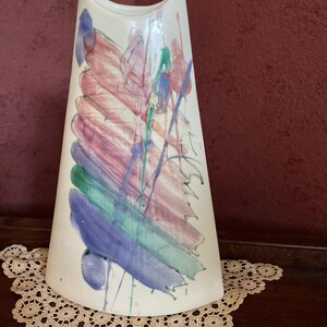 May include: A white ceramic vase with a flared, asymmetrical shape. The vase is decorated with colorful abstract brushstrokes in shades of red, purple, green, and blue. The vase is approximately 12 inches tall.