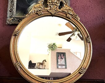 May include: A round, gold-framed mirror with ornate detailing around the edge. The mirror is reflecting a room with a ceiling fan and a picture on the wall.