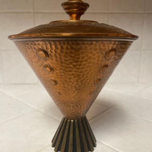 May include: A hammered copper vase with a conical shape and a decorative base. The vase has a lid with a finial on top. The vase is a mid-century modern design.