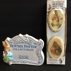 May include: A white ceramic sign with a Beatrix Potter® Collectibles logo and a Peter Rabbit illustration. The sign is next to a boxed set of two Beatrix Potter soap dishes featuring Peter Rabbit and Squirrel Nutkin.