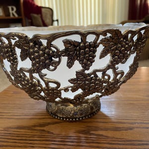 May include: A white frosted glass bowl with a silver metal grapevine design. The bowl is on a wooden table.