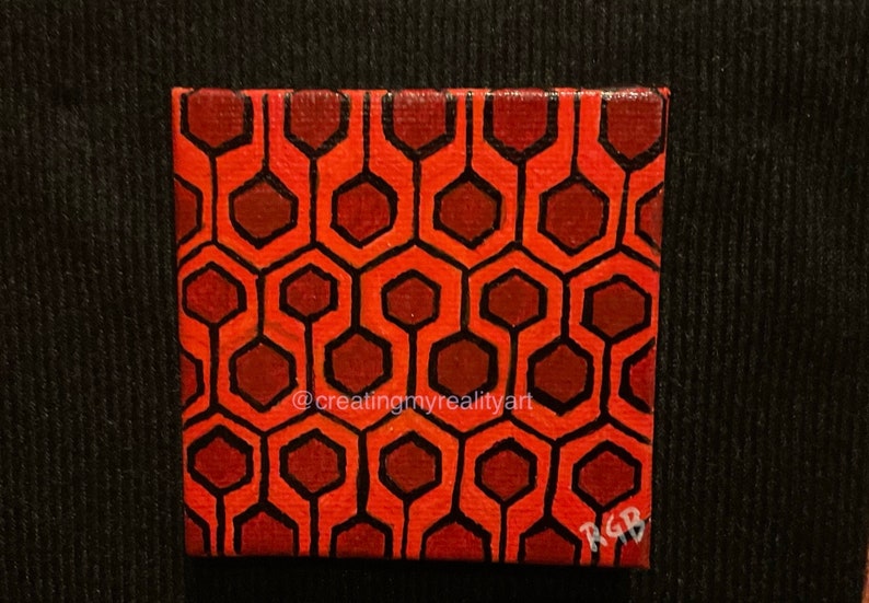 Puede incluir: Una peque&ntilde;a pintura sobre lienzo con un patr&oacute;n geom&eacute;trico rojo y marr&oacute;n. El patr&oacute;n es un dise&ntilde;o repetitivo de hex&aacute;gonos y l&iacute;neas. La pintura est&aacute; firmada "RGB" en la esquina inferior derecha. La pintura est&aacute; sobre un fondo negro.