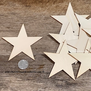 May include: A pile of 12 unfinished wooden stars of various sizes. The stars are arranged in a random pattern on a wooden surface. A single star is to the left of the pile, and a US quarter is next to it for scale.