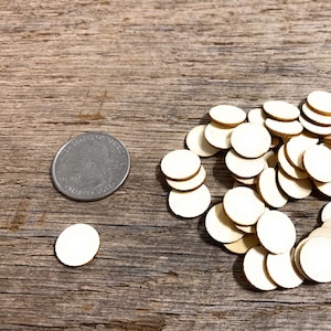 May include: A collection of small, round, unfinished wooden discs scattered on a weathered wooden surface. A U.S. quarter coin is placed nearby for size comparison. The discs are light beige in color and appear to be laser cut.