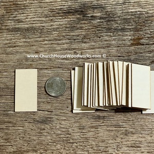 May include: A stack of 20 unfinished wood squares, each measuring approximately 1.5 inches by 2 inches. The squares are arranged in a neat pile, with the grain of the wood visible. A single square is placed to the left of the stack, along with a US quarter for scale.