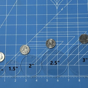 Może przedstawiać: Niebieska mata do cięcia z linijką oznaczoną w calach. Sześć przezroczystych plastikowych k&oacute;łek ułożonych jest w rzędzie, o średnicach 1 cala, 1,5 cala, 2 cale, 2,5 cala i 3 cale. Na macie leży pięć srebrnych monet, kt&oacute;re służą do pokazania rozmiaru k&oacute;łek.