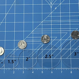 May include: A blue cutting mat with a ruler marked in inches. Six clear plastic circles are arranged in a row, with diameters of 1 inch, 1.5 inches, 2 inches, 2.5 inches, and 3 inches.  There are five silver coins on the mat, used to show the size of the circles.