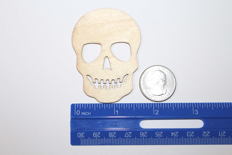 Pode incluir: Um recorte de madeira em forma de caveira, com aproximadamente 7,6 cm de altura, com &oacute;rbitas oculares, uma cavidade nasal e uma fileira de dentes. Uma moeda de um quarto de d&oacute;lar americano e uma r&eacute;gua azul est&atilde;o ao lado da caveira para compara&ccedil;&atilde;o de tamanho.