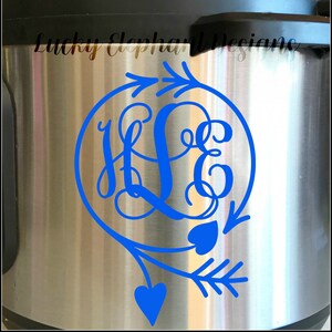 Puede incluir: Una calcomanía de monograma azul con las letras "HLE" en un círculo con flechas y corazones. La calcomanía está en una superficie de acero inoxidable.