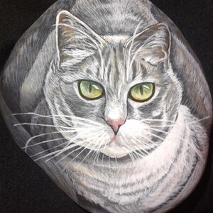 Puede incluir: Arte rupestre pintado a mano que representa un gato atigrado gris con ojos verdes. La cara del gato est&aacute; centrada en la piedra lisa de forma ovalada, mostrando pinceladas detalladas y sombreado realista. La obra de arte captura la expresi&oacute;n del gato con una nariz rosa y bigotes blancos.