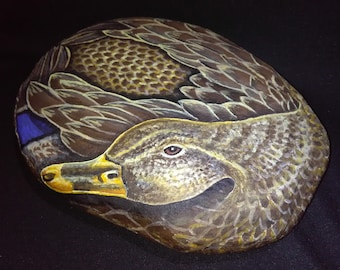 May include: Hand-painted rock art depicting a duck. The duck is painted in shades of brown, gray, and yellow, with a blue accent on the wing. The artwork is on a smooth, oval-shaped rock, showcasing detailed feather textures and a realistic eye.