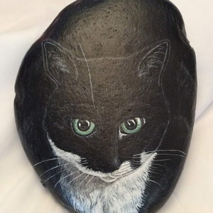 Puede incluir: Una roca negra pintada a mano con la cara de un gato con ojos verdes y marcas blancas. La cara del gato est&aacute; centrada en la roca, con bigotes blancos detallados y una mancha blanca en el pecho. La roca es lisa y de forma ovalada.