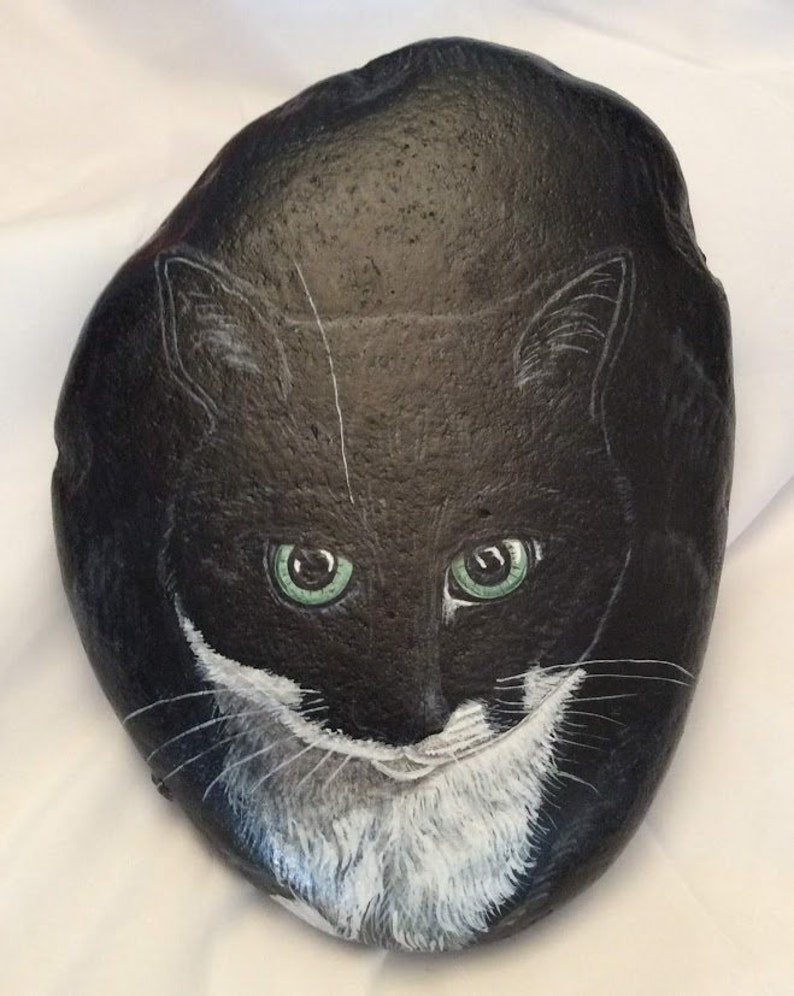 Puede incluir: Una roca negra pintada a mano con la cara de un gato con ojos verdes y marcas blancas. La cara del gato est&aacute; centrada en la roca, con bigotes blancos detallados y una mancha blanca en el pecho. La roca es lisa y de forma ovalada.