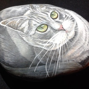 Puede incluir: Arte rupestre pintado a mano que representa un gato atigrado gris con ojos verdes. El pelaje del gato est&aacute; detallado con pinceladas blancas y grises, y sus ojos son de color verde brillante. La obra de arte est&aacute; sobre una roca lisa y ovalada.