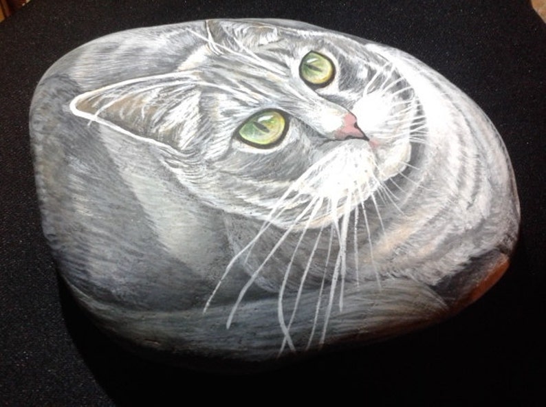 Puede incluir: Arte rupestre pintado a mano que representa un gato atigrado gris con ojos verdes. El pelaje del gato est&aacute; detallado con pinceladas blancas y grises, y sus ojos son de color verde brillante. La obra de arte est&aacute; sobre una roca lisa y ovalada.