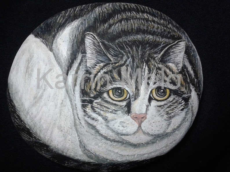 Puede incluir: Una piedra pintada a mano con un gato atigrado. El gato est&aacute; representado en tonos grises, blancos y marrones, con ojos amarillos y una nariz rosada. La obra de arte est&aacute; sobre un fondo negro. El nombre del artista, Karin Melo, es visible.
