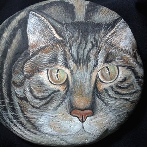 Puede incluir: Arte rupestre pintado a mano que representa la cara detallada de un gato atigrado. La obra de arte muestra los expresivos ojos verdes, la nariz rosa y el pelaje rayado del gato en tonos grises, marrones y negros. La cara del gato est&aacute; centrada en una piedra gris redondeada.