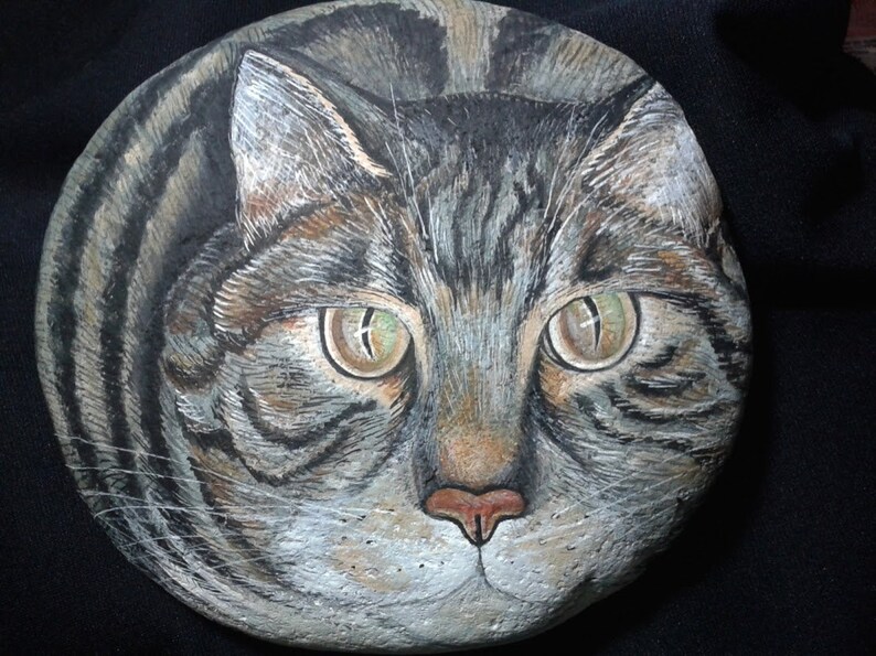 Puede incluir: Arte rupestre pintado a mano que representa la cara detallada de un gato atigrado. La obra de arte muestra los expresivos ojos verdes, la nariz rosa y el pelaje rayado del gato en tonos grises, marrones y negros. La cara del gato est&aacute; centrada en una piedra gris redondeada.