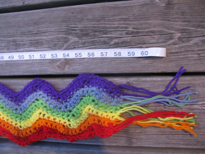 May include: A rainbow crochet scarf with fringe. The scarf is made with different colors of yarn, including purple, blue, green, yellow, orange, and red. The scarf is approximately 59 inches long.