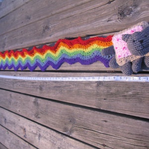 May include: A crocheted rainbow trail with a gray and pink crocheted cat figure at the end. The trail is made of different colored yarn, including red, orange, yellow, green, blue, and purple. The cat figure has a pink heart on its chest and is sitting on a wooden surface.