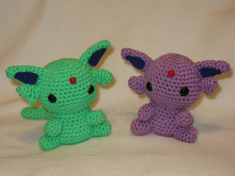 May include: Two crocheted Pokemon plushies. The one on the left is green with blue ears and black eyes. The one on the right is purple with blue ears and black eyes.