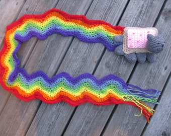 Puede incluir: Una bufanda de crochet arcoíris con un gato de crochet gris que lleva un disfraz de pan rosa y marrón. La bufanda está sobre una superficie de madera.