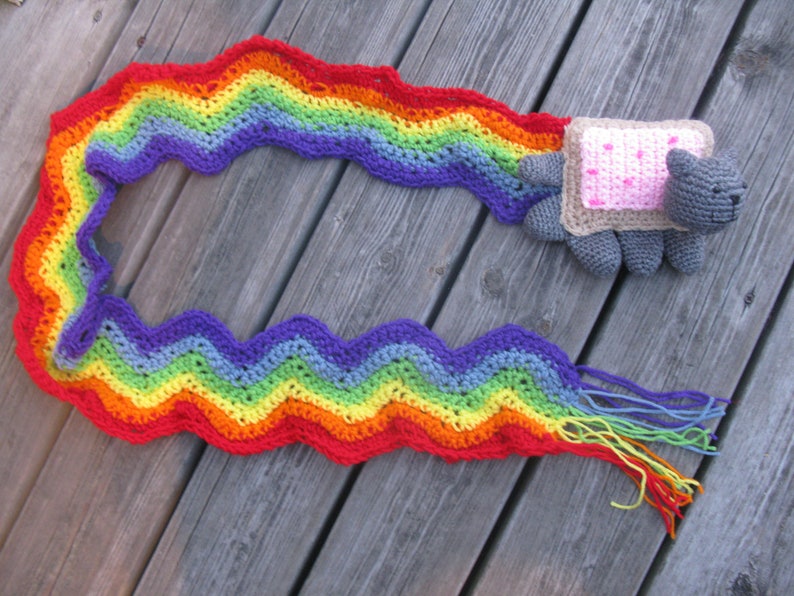 May include: A rainbow crocheted scarf with a gray crocheted cat wearing a pink and brown bread costume. The scarf is laying on a wooden surface.