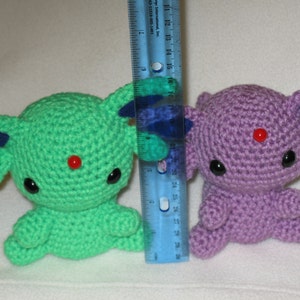 May include: Two crocheted plushies, one green and one purple, with red bead eyes and red noses. The plushies are sitting on a white surface next to a ruler.
