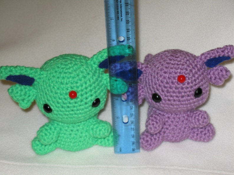May include: Two crocheted plushies, one green and one purple, with red bead eyes and red noses. The plushies are sitting on a white surface next to a ruler.