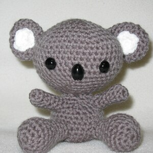 May include: A crocheted koala bear in grey. The koala has large black eyes, a black nose, and white accents inside its ears. The arms and legs are small and rounded, giving it a cuddly appearance. A handmade soft toy, ideal for children.