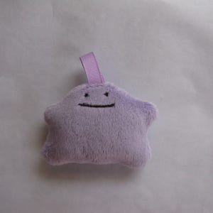 May include: A purple plush toy shaped like a star with a black smiling face. The toy has a purple ribbon attached to the top.