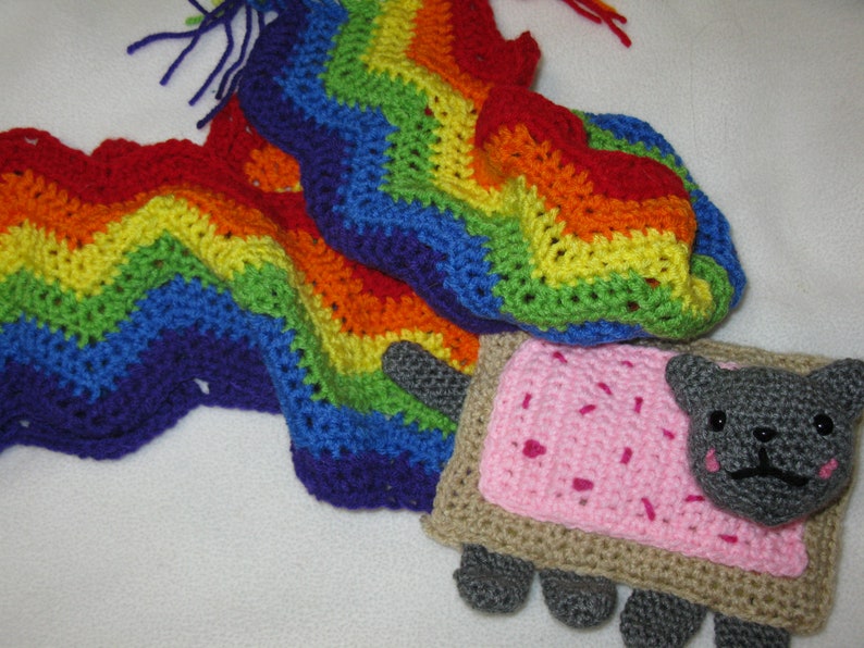 May include: A crocheted rainbow blanket with a crocheted 'Nyan Cat' toy. The cat is gray with pink frosting and sprinkles on its back.