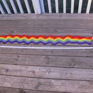 May include: A long, rainbow-colored crocheted scarf with a wavy pattern. The scarf is laid out on a wooden surface and measures approximately 5 feet in length.