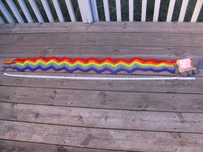 May include: A long, rainbow-colored crocheted scarf with a wavy pattern. The scarf is laid out on a wooden surface and measures approximately 5 feet in length.